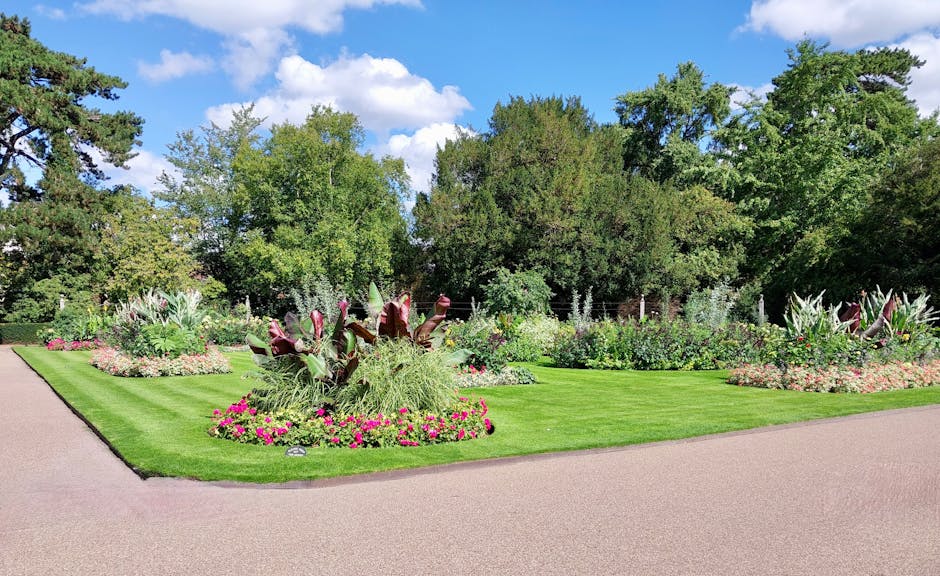 A well-maintained formal garden with a neatly mown green lawn bordered by ornamental flower beds featuring vibrant pink, purple, and white blooms. The flower beds include a variety of foliage plants w