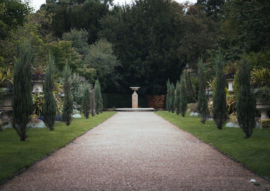 A well-maintained formal garden with a neatly mown green lawn bordered by ornamental flower beds featuring vibrant pink, purple, and white blooms. The flower beds include a variety of foliage plants w